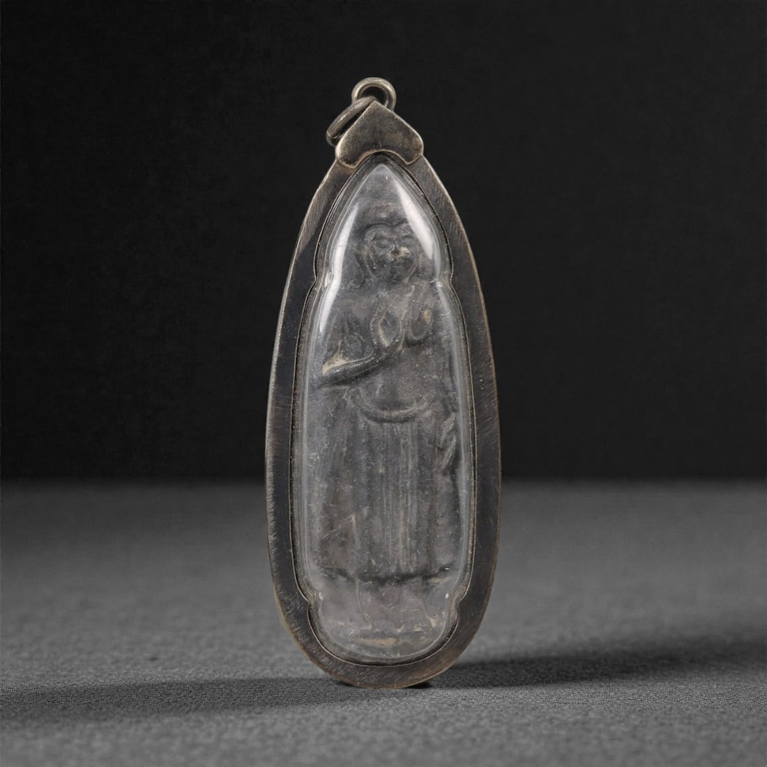 A Thai Standing Buddha Amulet with Metal Frame, 20th Century, 28FP: The amulet is of elongated, teardrop-shaped form, featuring a molded standing Buddha figure on the obverse, rendered in a dark patinated material against a light beige ground. The Buddha is depicted w