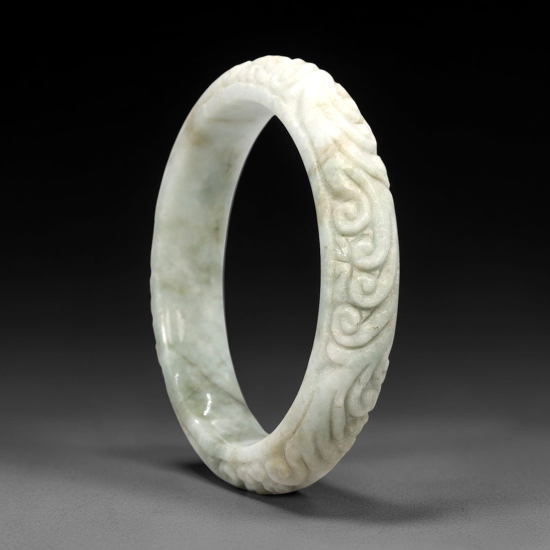 A Chinese Jadeite Jade Bangle Bracelet, 20th Century, 46FS: Carved from pale celadon jadeite jade with natural tonal variation and subtle russet inclusions, the bangle is formed as a continuous circular band. The exterior surface is decorated with raised relie