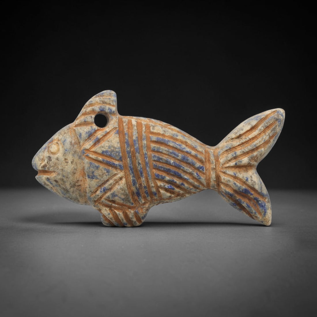 Ancient Lapis Fish Amulet - Near Eastern Carved Figurine, 63ZX: Carved from lapis lazuli, the amulet takes the form of a stylized fish, rendered in a flat, relief style with incised linear decoration. The body is segmented with incised lines to represent scales an