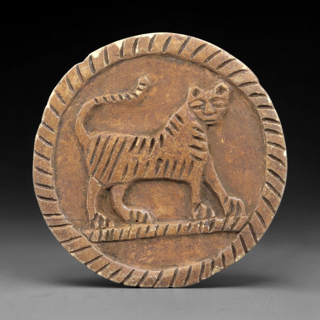 Ancient Steatite Plaque - Near Eastern Feline Relief, 90BE: Carved from steatite, the circular plaque features a raised relief of a striding feline, likely a tiger or lion, rendered with incised linear details defining its striped body, curled tail, and facial