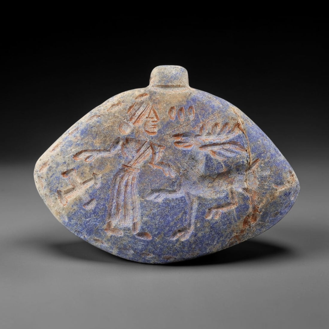 Ancient Lapis Amulet - Near Eastern Figural Animal Scene, 79BF: Carved from lapis lazuli, the amulet is a rounded, shield-shaped plaque with a perforated suspension knob at the upper edge, designed for wearable use. The front face is incised with a dynamic figural
