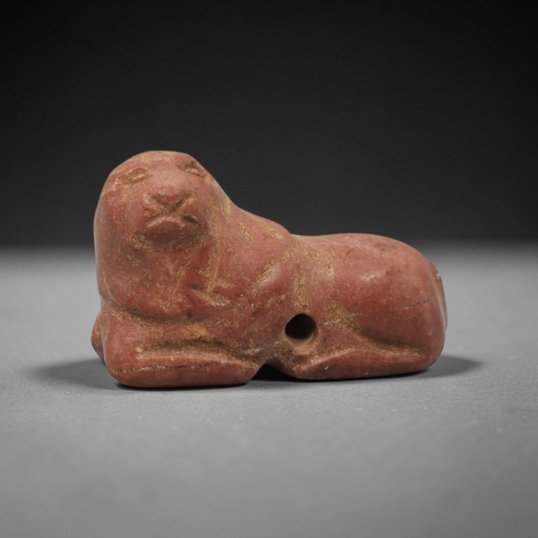 Ancient Stone Feline Amulet - Near Eastern Figurine, 88XV: This amulet is carved from a warm reddish stone with natural earthy patina and subtle tonal variations across its surface, executed in a simplified, stylized manner that emphasizes the recumbent felin