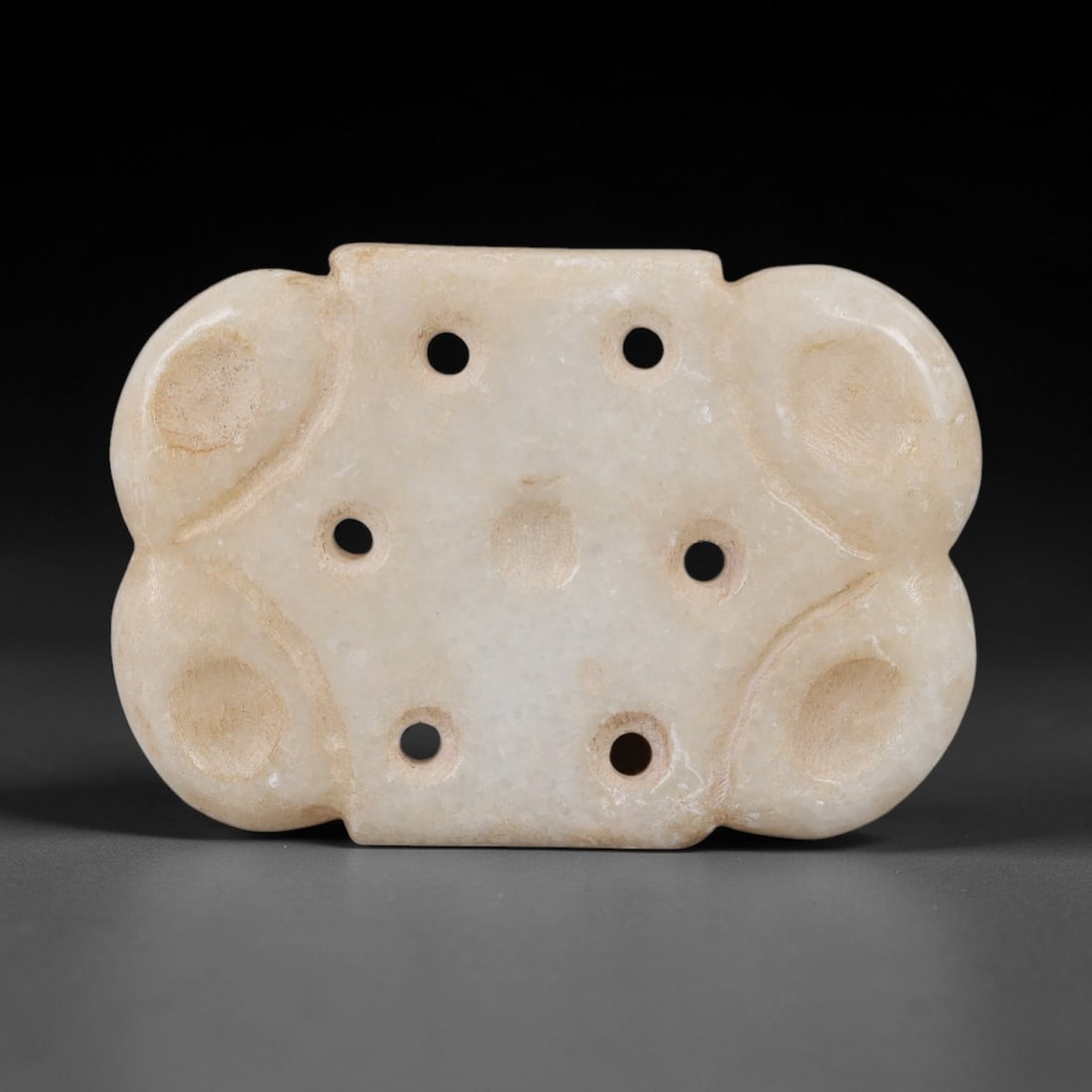 Ancient Marble Amulet - Near Eastern Ritual Pendant, 18AK: Crafted from pale marble, this amulet features a symmetrical, lobed rectangular form with rounded lateral projections. The surface is punctuated by six circular suspension holes arranged in two horizo