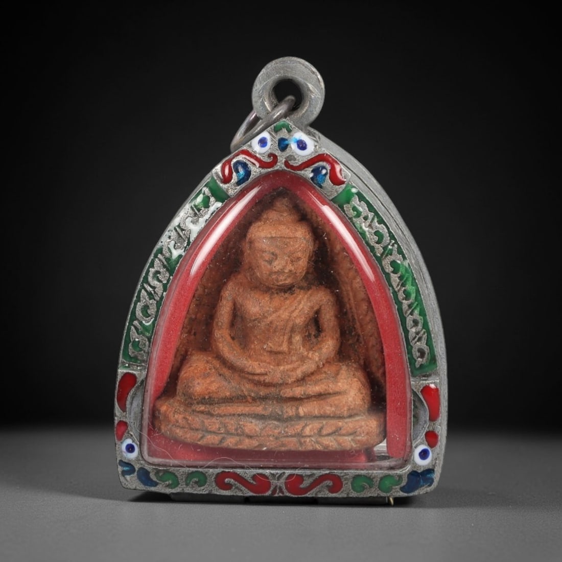 A Thai Terracotta Buddha Amulet in an Enameled Silver Frame, 20th Century, 84ZL: The amulet depicts the Buddha seated in meditative pose atop a lotus base, cast in terracotta with a warm, earthy tone and visible surface texture. The figure is encased in a clear protective cover wi