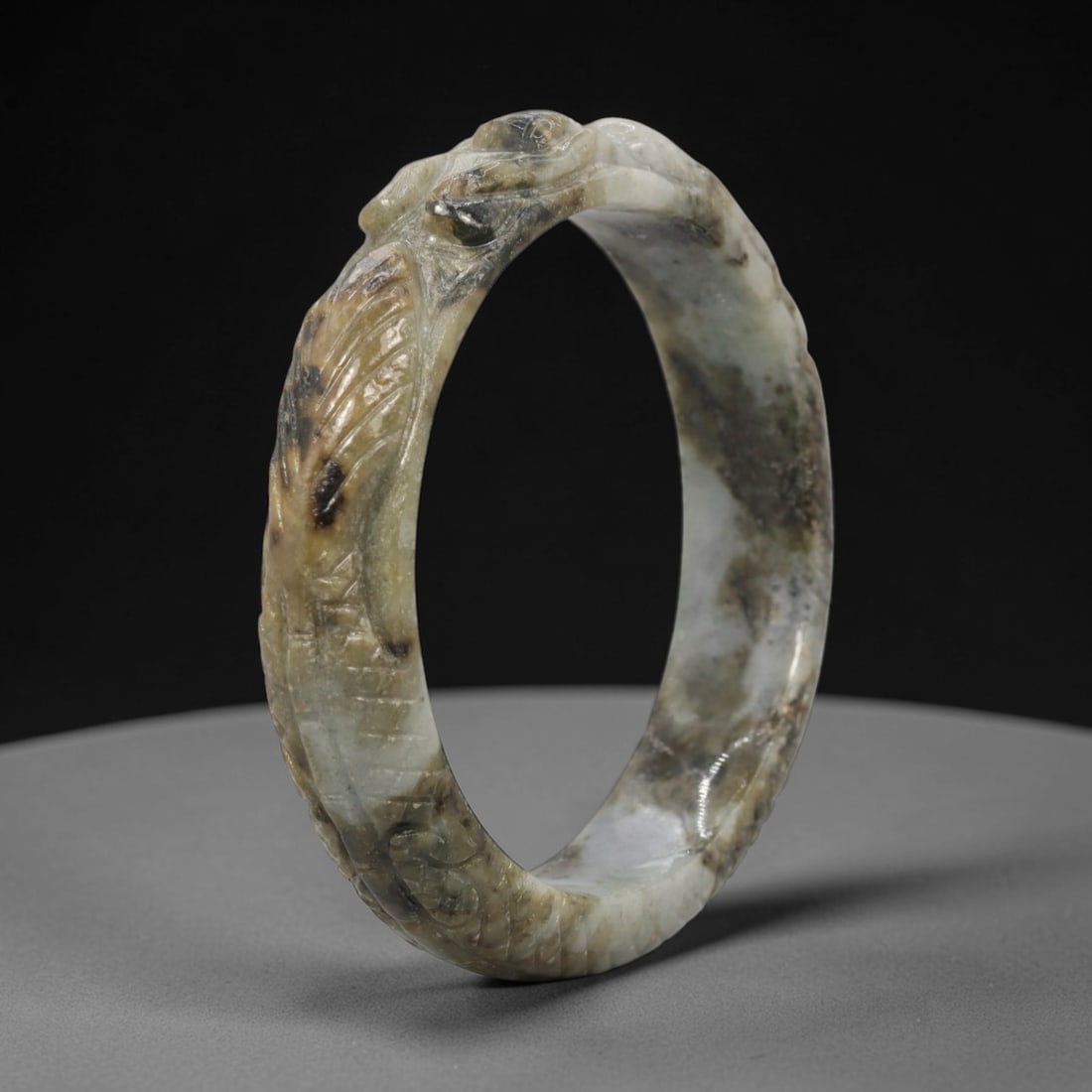 A Chinese Carved Jadeite Jade Bangle Bracelet, 20th Century, 70ZL: A fine jadeite bangle of circular form, masterfully carved in relief with a continuous band of a sinuous dragon coiling around its circumference. The stone exhibits a striking variegated palette, with