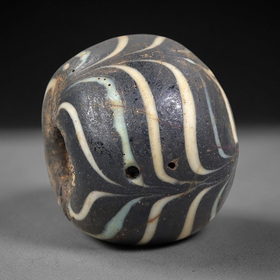 Ancient Glass Bead - Central Asian Islamic Amulet, 42BF: A barrel-shaped glass bead crafted from dark core glass, decorated with applied trailing in opaque white and pale blue, arranged in a flowing, radiating wave pattern across the surface. The bead featu