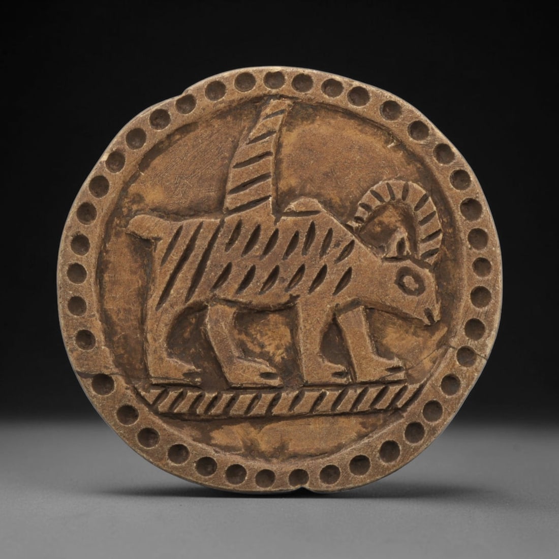 Ancient Steatite Plaque - Near Eastern Winged Ram Relief, 26ZY: Carved from steatite, this circular plaque features a relief depiction of a stylized winged ram, rendered in the schematic artistic tradition of the Ancient Near East. The creature is shown walking in