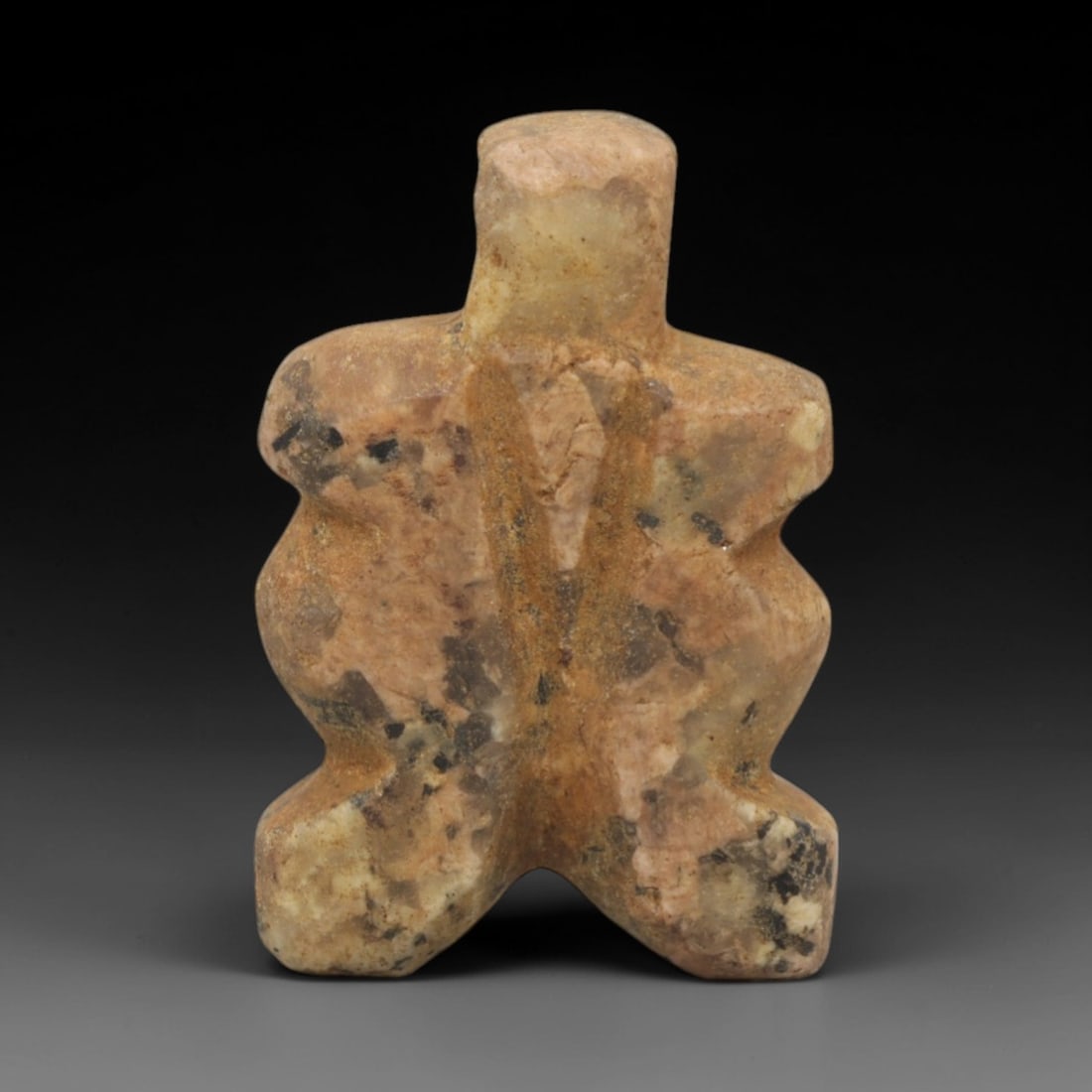 Ancient Stone Crossroad Idol - Near Eastern Amulet, 6XR: Carved from a mottled ochre and black stone, this amulet takes a compact, cross-shaped form with a cylindrical head, broad angular shoulders, and splayed legs forming a cross-like base. The geometric,