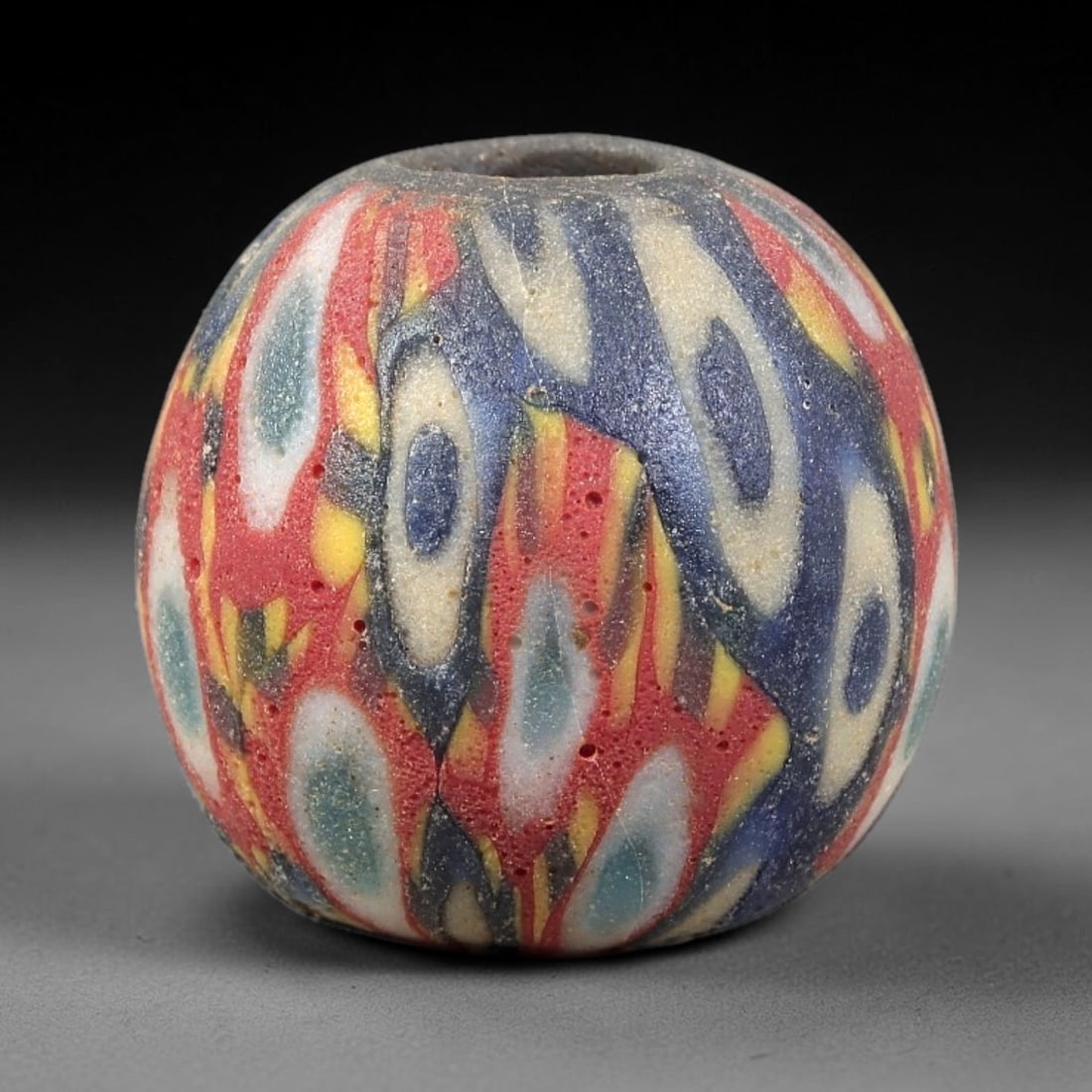 Ancient Glass Bead - Roman Millefiori Spherical Pendant, 38BG: A spherical ancient glass bead crafted in the millefiori mosaic technique, with a vibrant multi-layered cane pattern across its surface. The bead features interwoven oval motifs in red, blue, white, y