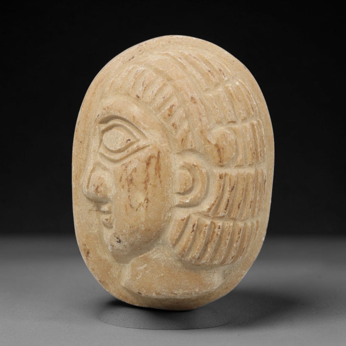 Ancient Stone Amulet - Near Eastern Bust Plaque Pendant, 9ZX: Carved from light-colored stone, the amulet is an oval plaque crafted in the ancient Near Eastern style, featuring a raised relief portrait of a human bust in profile. The surface bears incised detail