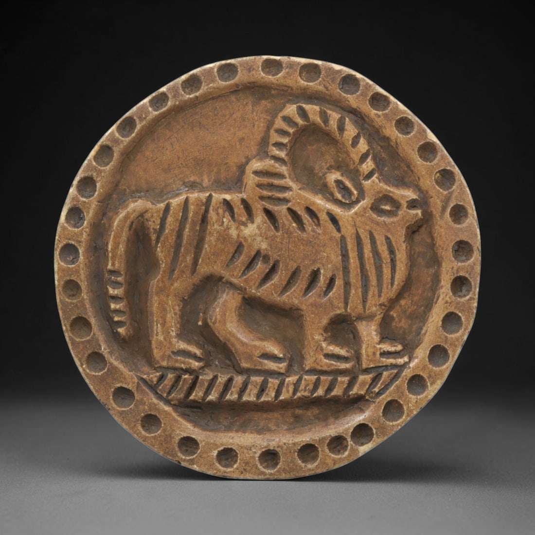 Ancient Steatite Plaque - Near Eastern Ram Relief, 23ZY: Carved from steatite, this circular plaque features a relief depiction of a stylized ram, rendered in the schematic artistic tradition of the Ancient Near East. The animal is shown standing in profile