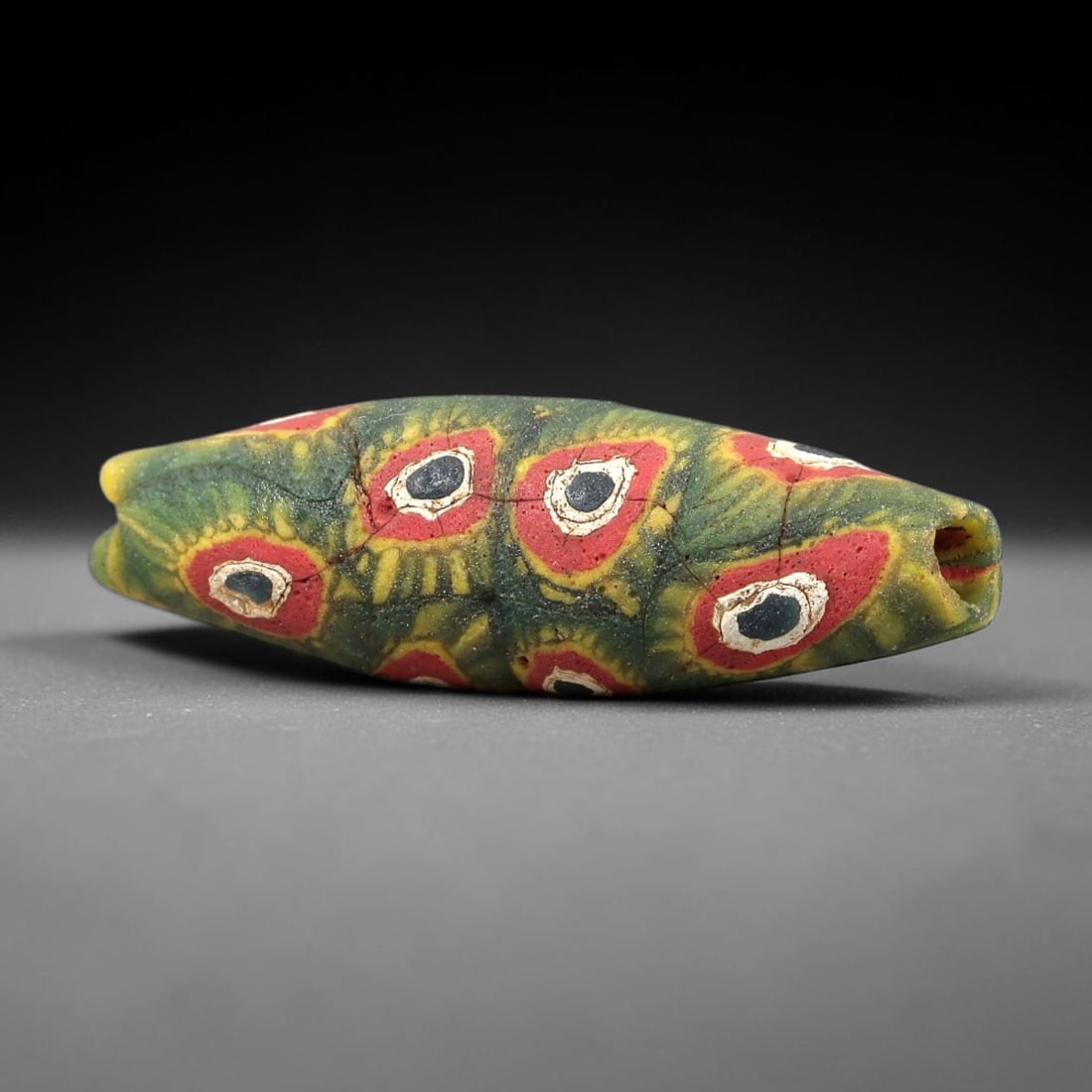 Ancient Glass Bead - Roman Millefiori Elongated Eye Bead Pendant, 18BG: An elongated, bicone-shaped ancient glass bead crafted in the millefiori mosaic technique, with a vibrant green glass base overlaid with intricate fused cane patterns. The surface is decorated with re