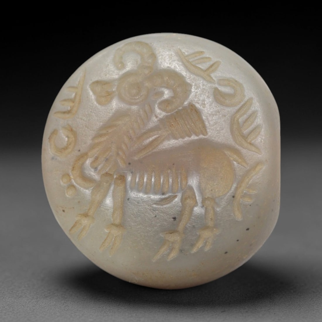 Ancient Chalcedony Hemispherical Stamp Seal - Near Eastern Ram & Script, 35ZP: Crafted from translucent, pale chalcedony, this hemispherical stamp seal bears a deeply incised depiction of a stylized ram, rendered in linear relief with distinct curved horns, patterned torso, and 