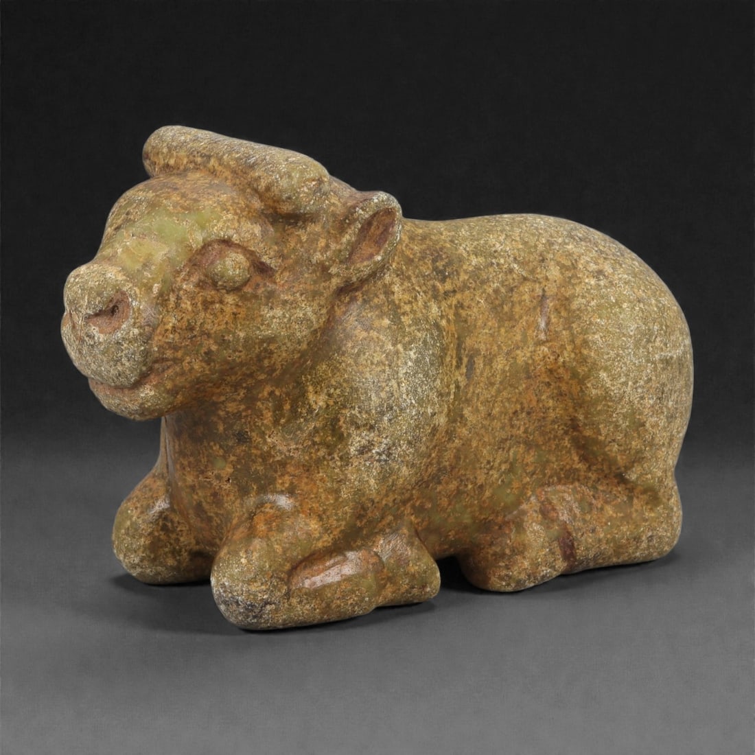 An Achaemenid Serpentine Bovid Figurine, 48ZB: Carved from mottled greenish-brown serpentine with areas of surface patination, the figurine depicts a recumbent bovid with the forelegs folded beneath the chest and the hindquarters compactly positio