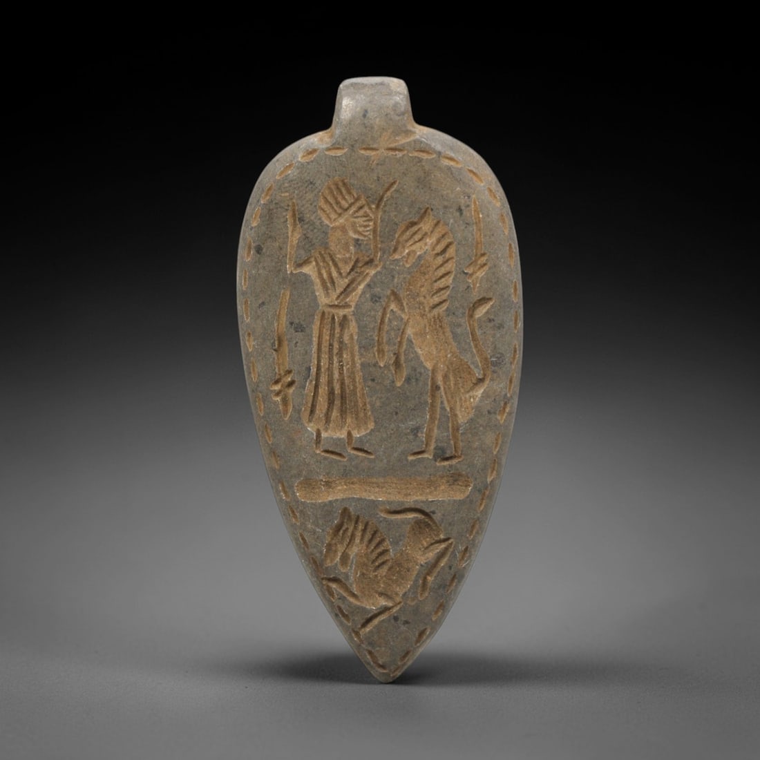 Ancient Near Eastern Stone Figural Amulet - Pendant, 38ZR: This artifact is a carved stone figural amulet pendant, crafted from a fine-grained, grey-brown stone with a matte surface and earthy patina filling the incised details. The teardrop-shaped body featu