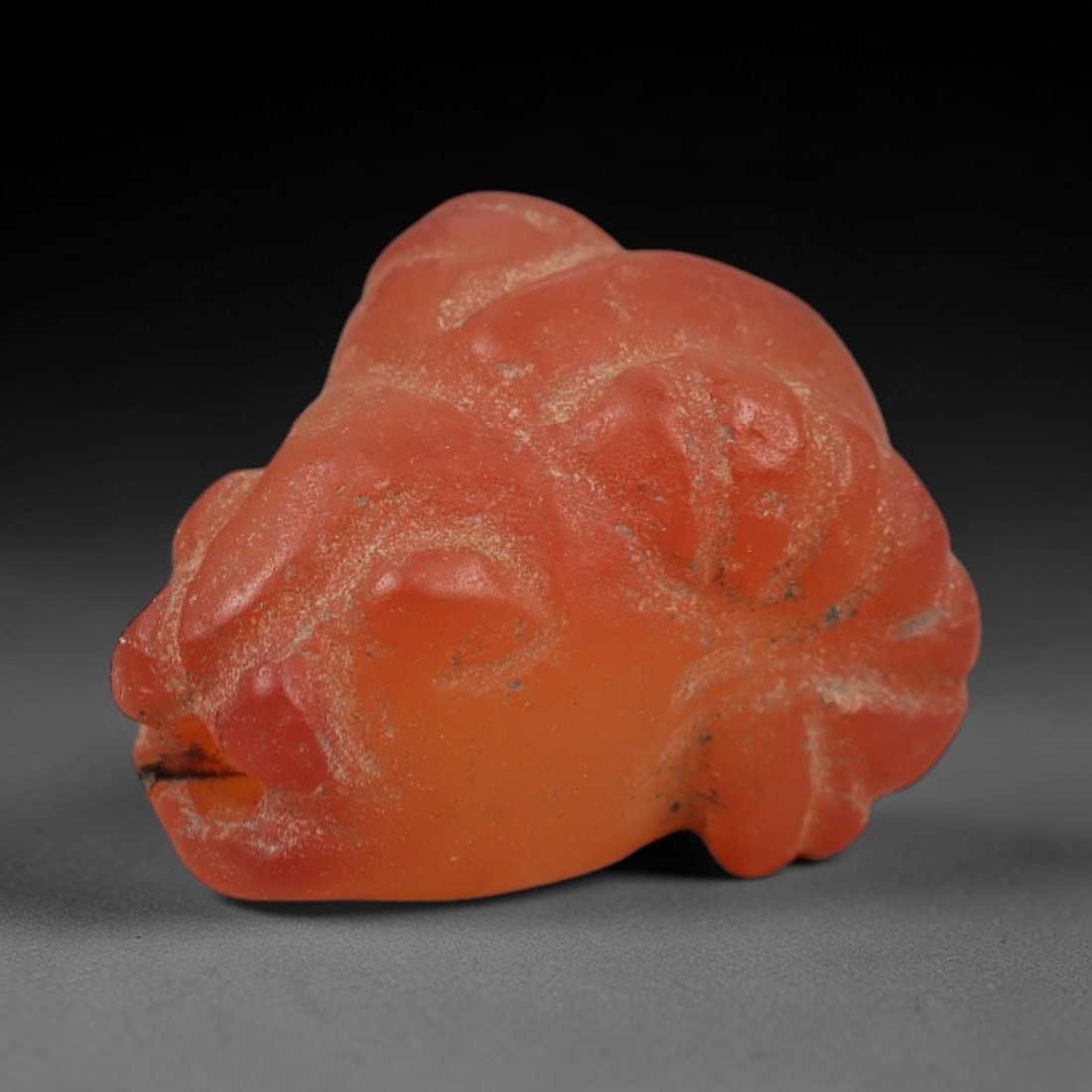 Ancient Carnelian Ram's Head Amulet - Near Eastern Figurine, 54XS: Carved from warm, translucent orange-red carnelian with natural tonal gradients and subtle surface patination, this amulet is formed as a stylized ram's head with incised linear detailing to define fa