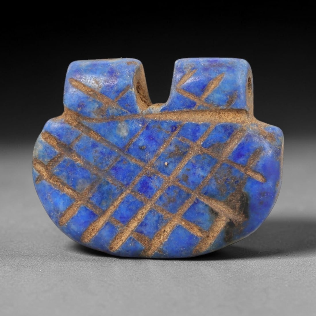 Ancient Lapis Lazuli Amulet - Egyptian Grid Pattern Pendant, 19AP: Carved from lapis lazuli, this amulet features a rounded, shield-like form with two raised upper lobes framing a central suspension channel, its surface incised with a precise grid pattern of intersec