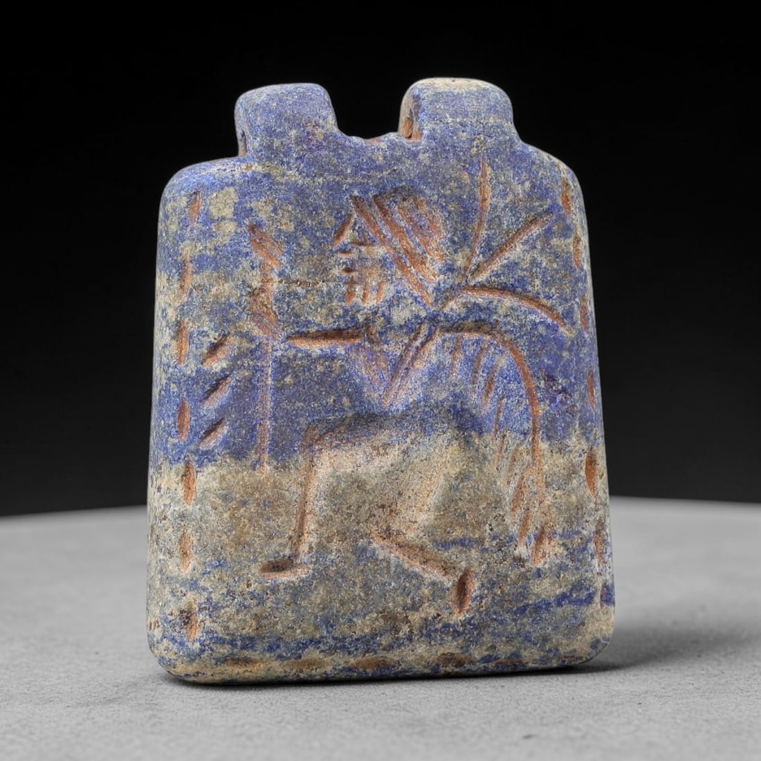 Ancient Lapis Amulet - Near Eastern Figural Pendant, 82BF: Carved from lapis lazuli, the amulet is a rectangular plaque with two perforated suspension knobs at the top, designed for wearable use. The front face is incised with a dynamic, stylized humanoid fig