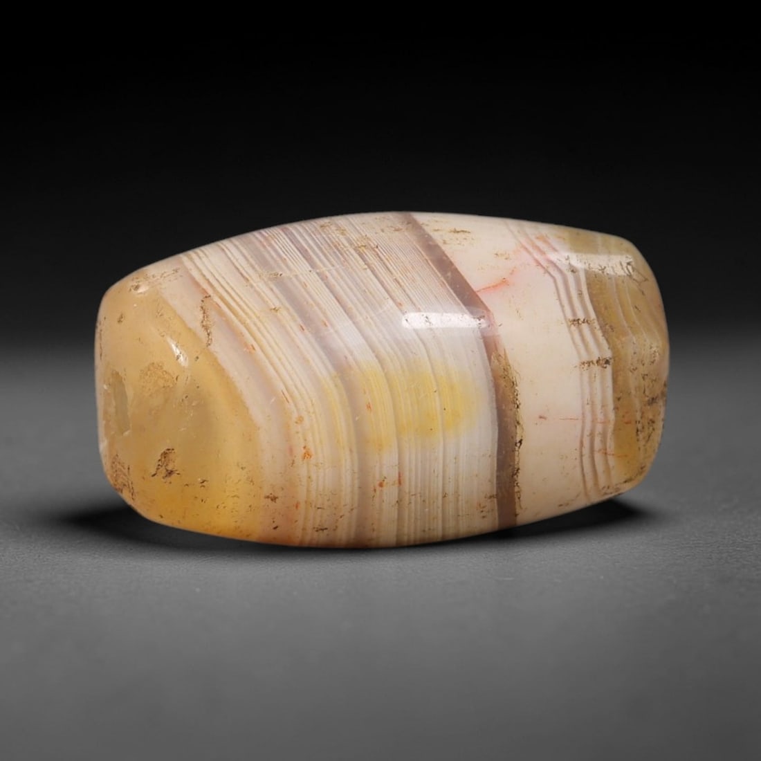 Ancient Agate Bead - Near Eastern Amulet, 77BC: This artifact is crafted from banded agate, exhibiting fine concentric layers of warm honey, creamy white, and soft grey tones, shaped into a smooth barrel form through skilled ancient lapidary work. 