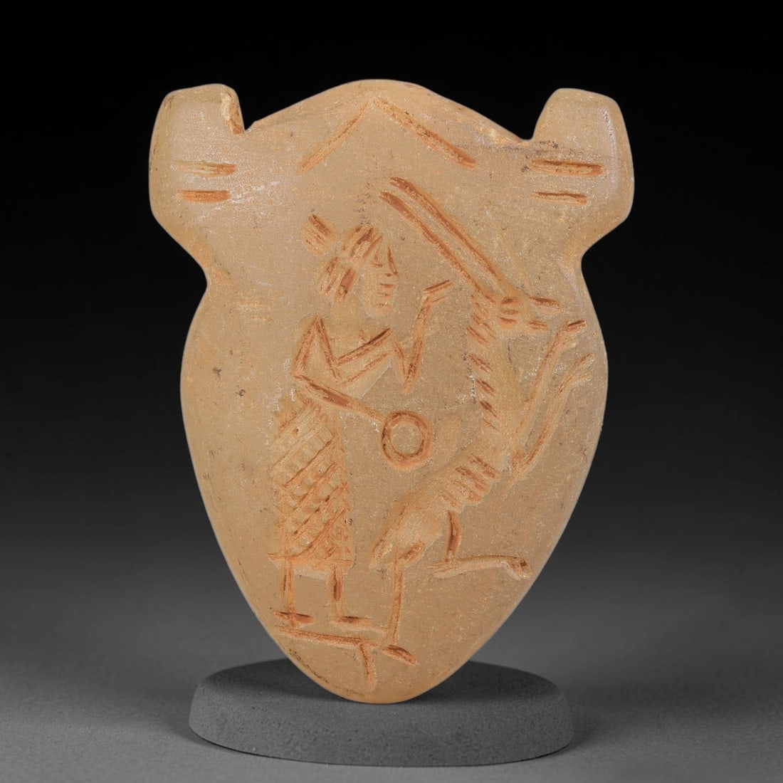 Ancient Stone Hunter & Ibex Amulet - Near Eastern Carved Pendant, 26BA: Carved from translucent light-colored hardstone, the shield-shaped amulet features a relief-carved figural scene, rendered with incised detail and filled with red pigment for contrast. The composition