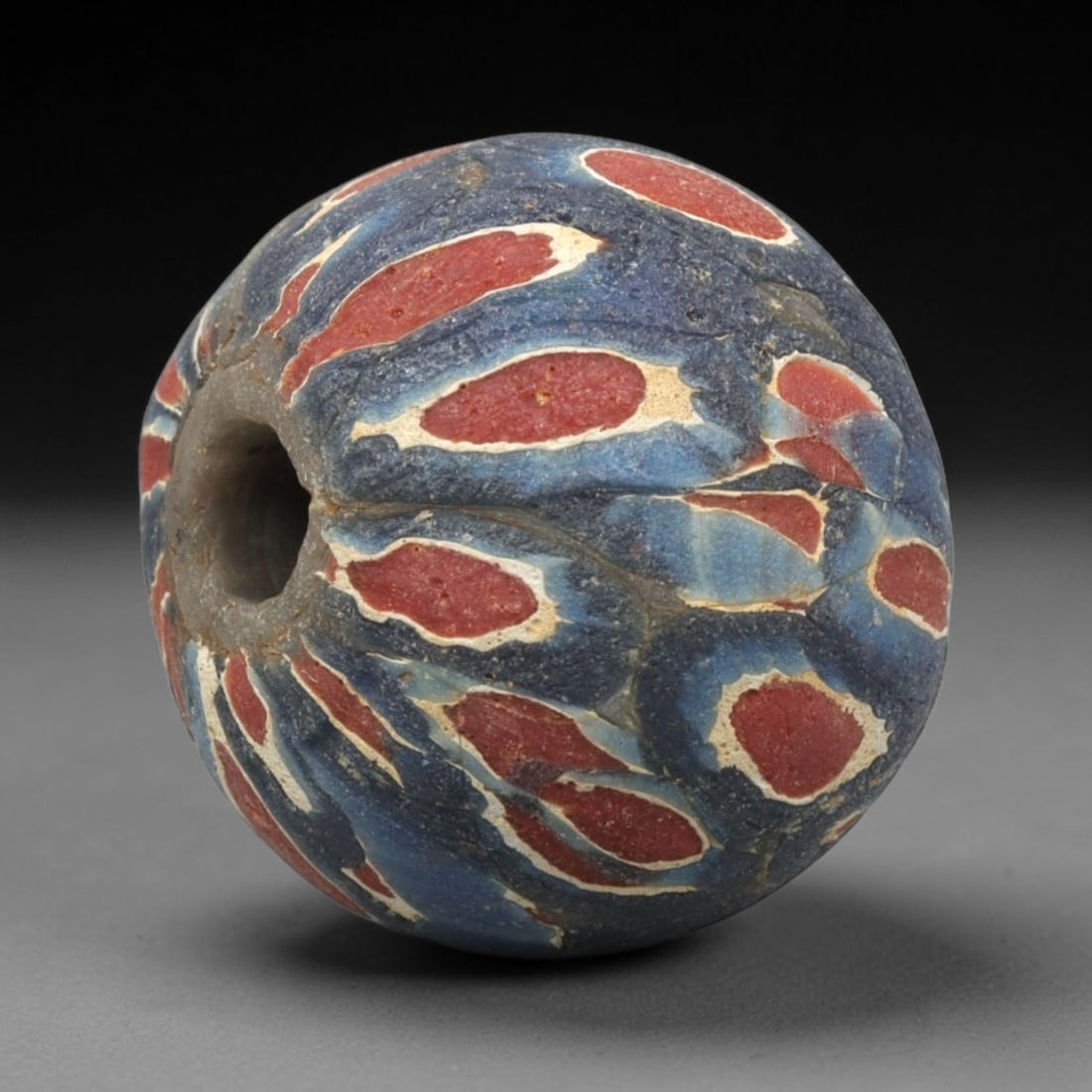 Ancient Mosaic Glass Bead - Roman Egyptian Floral Amulet, 62WD: This spherical bead is crafted from core-formed mosaic glass, featuring a deep blue base decorated with repeating teardrop-shaped floral motifs. Each motif consists of a central red glass inlay outlin