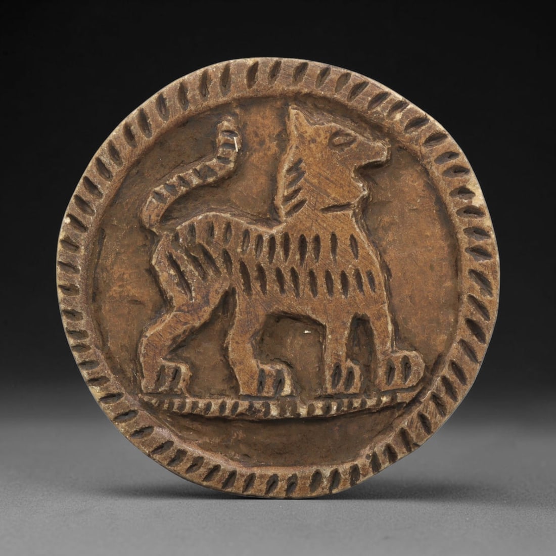 Ancient Steatite Plaque - Near Eastern Canine Relief, 35ZY: Carved from steatite, this circular plaque presents a relief carving of a canine figure, depicted in a schematic yet expressive style. The animal stands in profile, with incised linear details articul