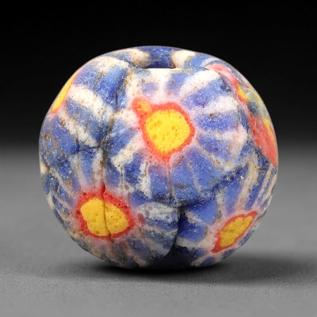 Ancient Glass Bead - Roman Millefiori Floral Pendant, 33BG: A spherical ancient glass bead crafted in the millefiori mosaic technique, with a deep blue glass base overlaid with intricate fused cane patterns. The surface is decorated with repeating floral motif
