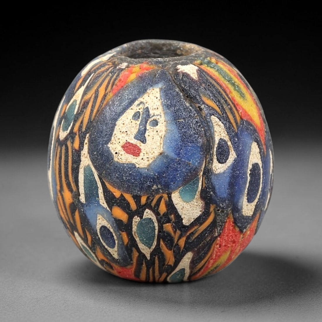 Ancient Glass Bead - Roman Millefiori Face Pendant, 21BG: A spherical ancient glass bead crafted in the millefiori mosaic technique, with a complex multi-layered cane pattern featuring a central human face motif. The face is rendered in white glass with blue