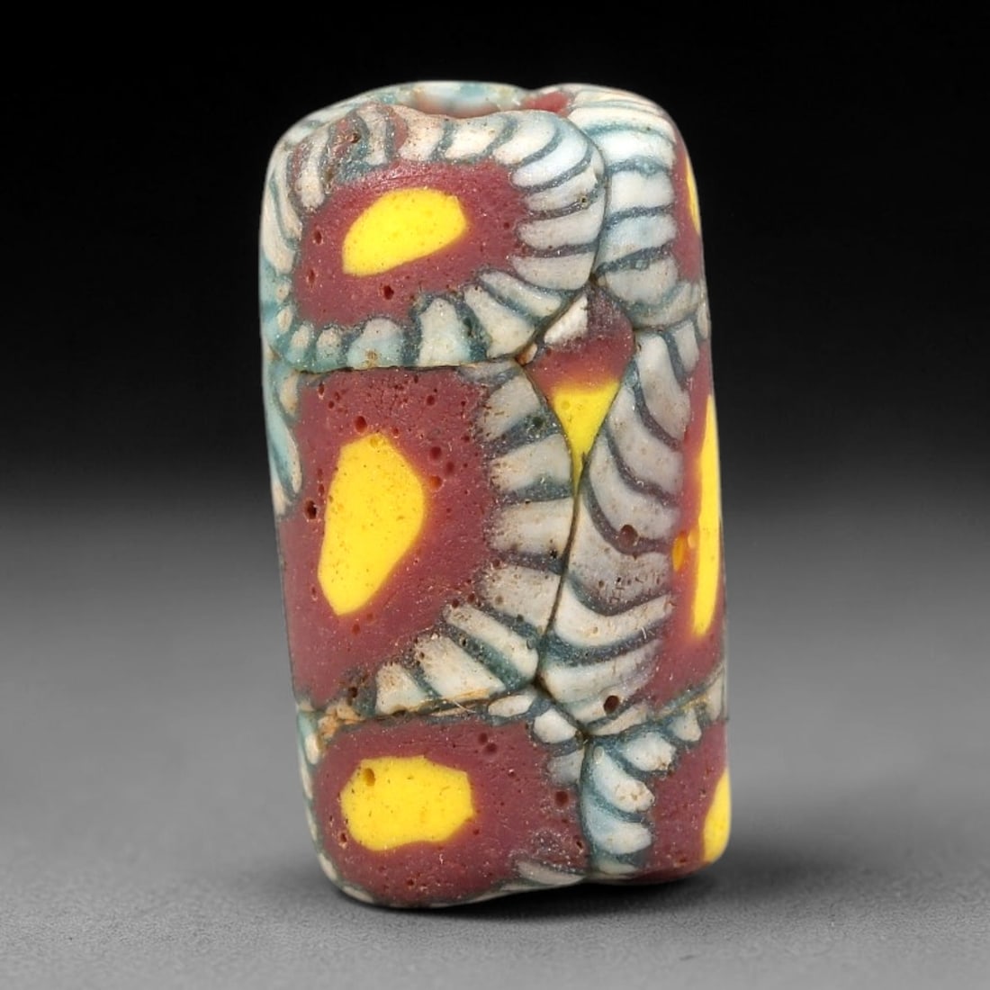 Ancient Glass Bead - Roman Millefiori Cylindrical Pendant, 19BG: A cylindrical ancient glass bead crafted in the millefiori mosaic technique, constructed from fused glass canes arranged in a complex floral and geometric pattern. The surface features repeating sunbu