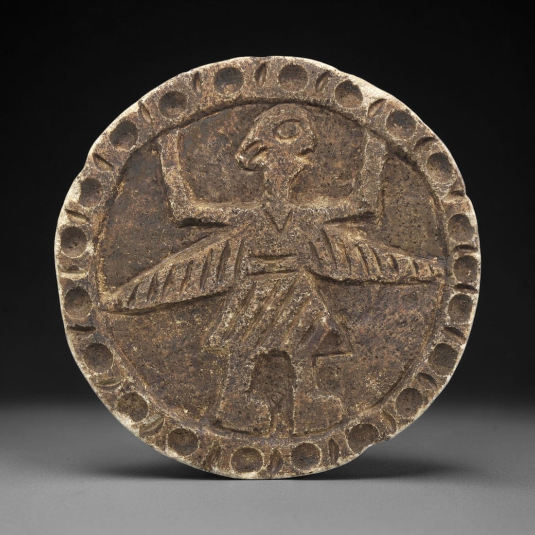 Ancient Steatite Plaque - Near Eastern Winged Figure Relief, 19ZY: Carved from steatite, this circular plaque features a relief depiction of a winged anthropomorphic figure, rendered in the stylized, schematic artistic tradition of the Ancient Near East. The figure s
