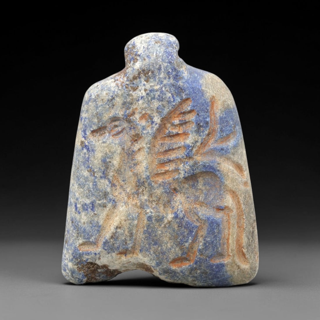 Ancient Lapis Amulet - Near Eastern Winged Creature, 78ZY: Carved from lapis lazuli, the amulet is incised with a dynamic winged quadrupedal creature, rendered in linear relief with pigmented detailing to accentuate form. The figure displays a stylized avian 