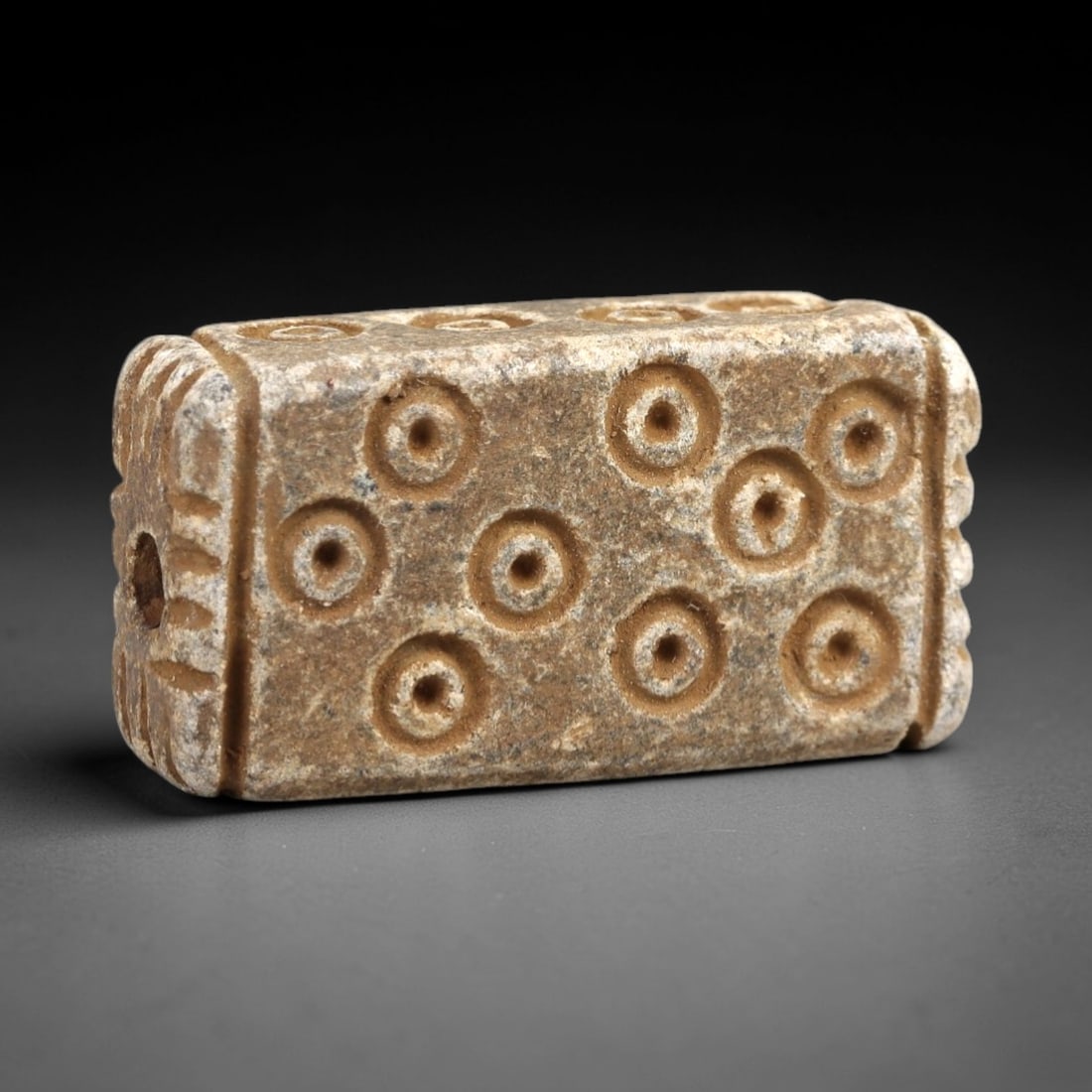 Ancient Steatite Amulet - Near Eastern Concentric Circles, 38AK: Crafted from steatite, this rectangular amulet features incised concentric circle motifs across its primary face, with decorative linear carvings framing the suspension hole on one lateral edge. The p