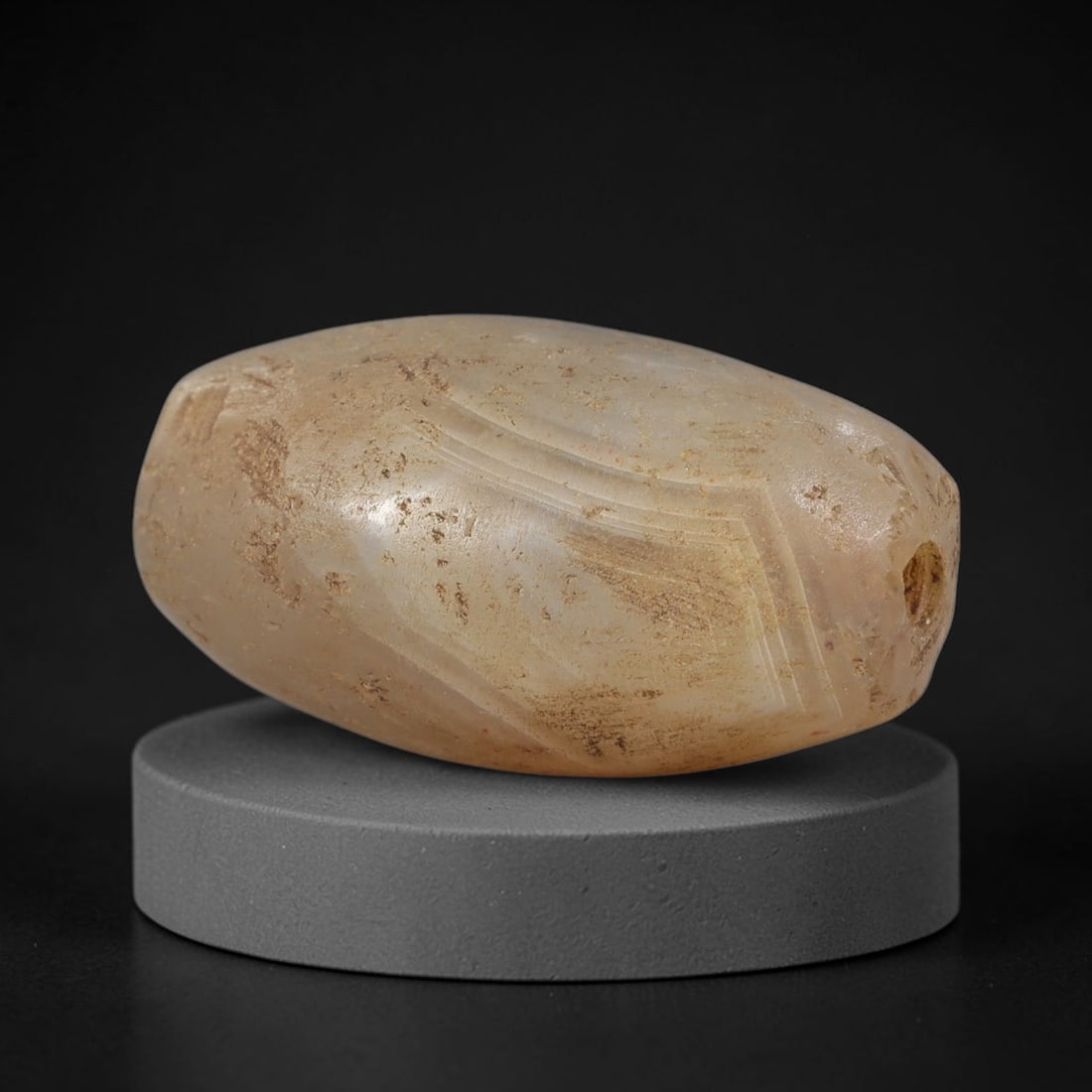 Ancient Agate Bead - Near Eastern Carved Artifact, 13BD: This artifact is carved from banded agate, shaped into an elongated ovoid form with incised chevron-style linear decoration across its surface. The bead features a central perforation for suspension,
