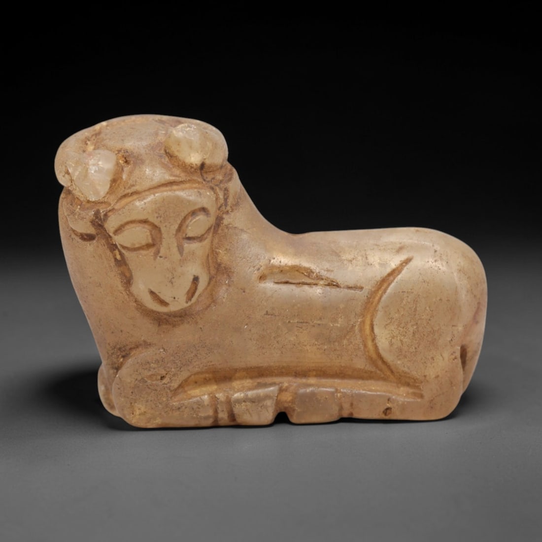 Ancient Rock Crystal Bovid - Near Eastern Amulet Figurine, 100ZP: Carved from translucent pale rock crystal, this reclining bovid figurine features stylized curved horns, closed incised eyes, and simplified facial features. The compact, rounded body is defined by su