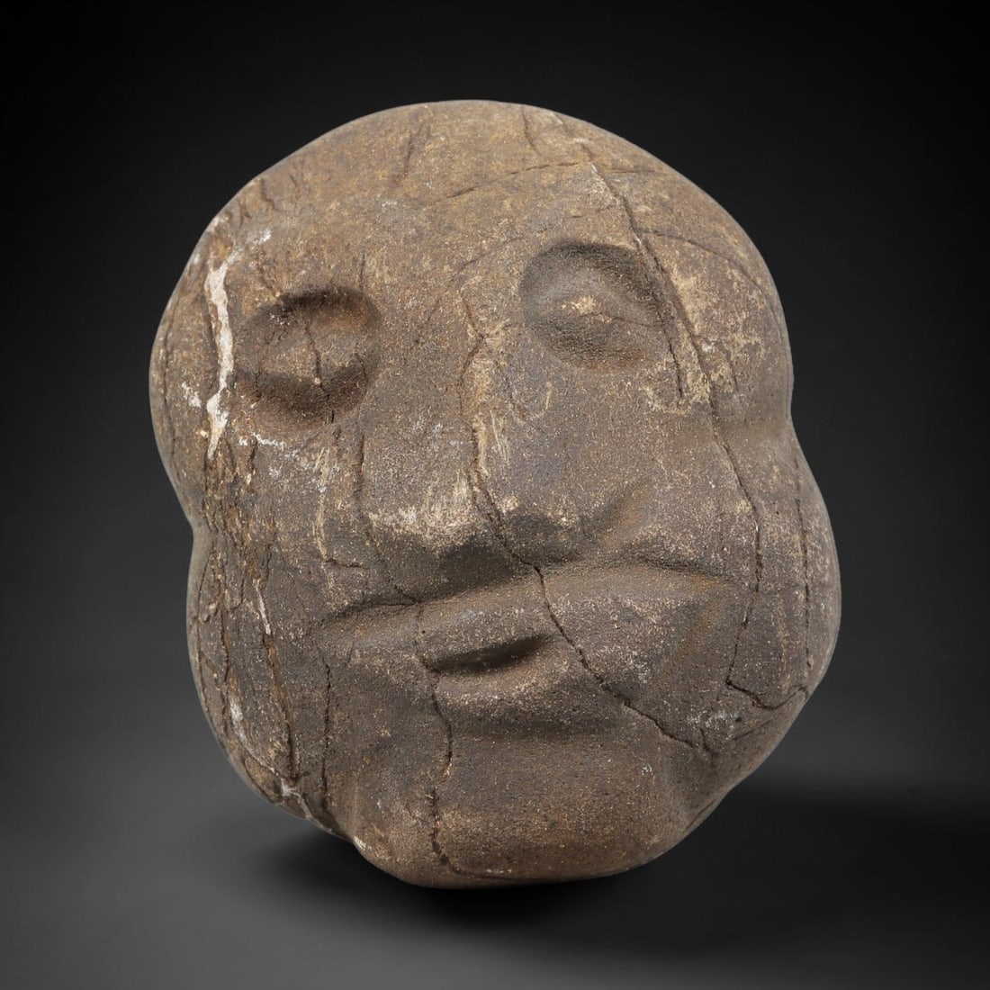 Ancient Stone Maskette - Near Eastern Anthropomorphic Head, 45WJ: Carved from a coarse, porous stone, this artifact takes the form of a stylized maskette or head fragment, rendered with simplified, schematic facial features. The eyes are carved as recessed, almond-s