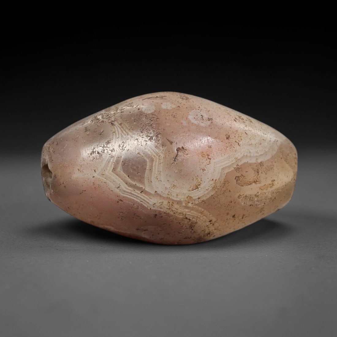 Ancient Agate Bead - Bactrian Amulet, 41BC: This artifact is fashioned from banded agate, showcasing intricate concentric layers of pale pinkish-brown and creamy white agate with subtle greyish mineral inclusions, shaped into a rounded barrel f