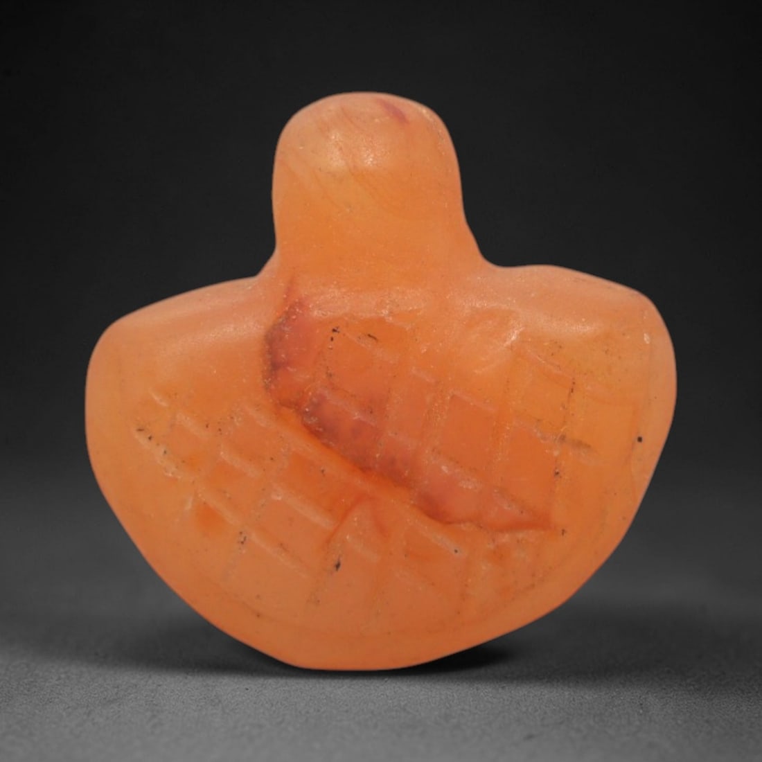 Ancient Carnelian Heart Amulet - Egyptian Figurine, 74XS: This amulet is carved from warm orange-red carnelian, shaped into a stylized heart form with incised crisscross patterning across its surface, characteristic of Egyptian amuletic iconography. The comp