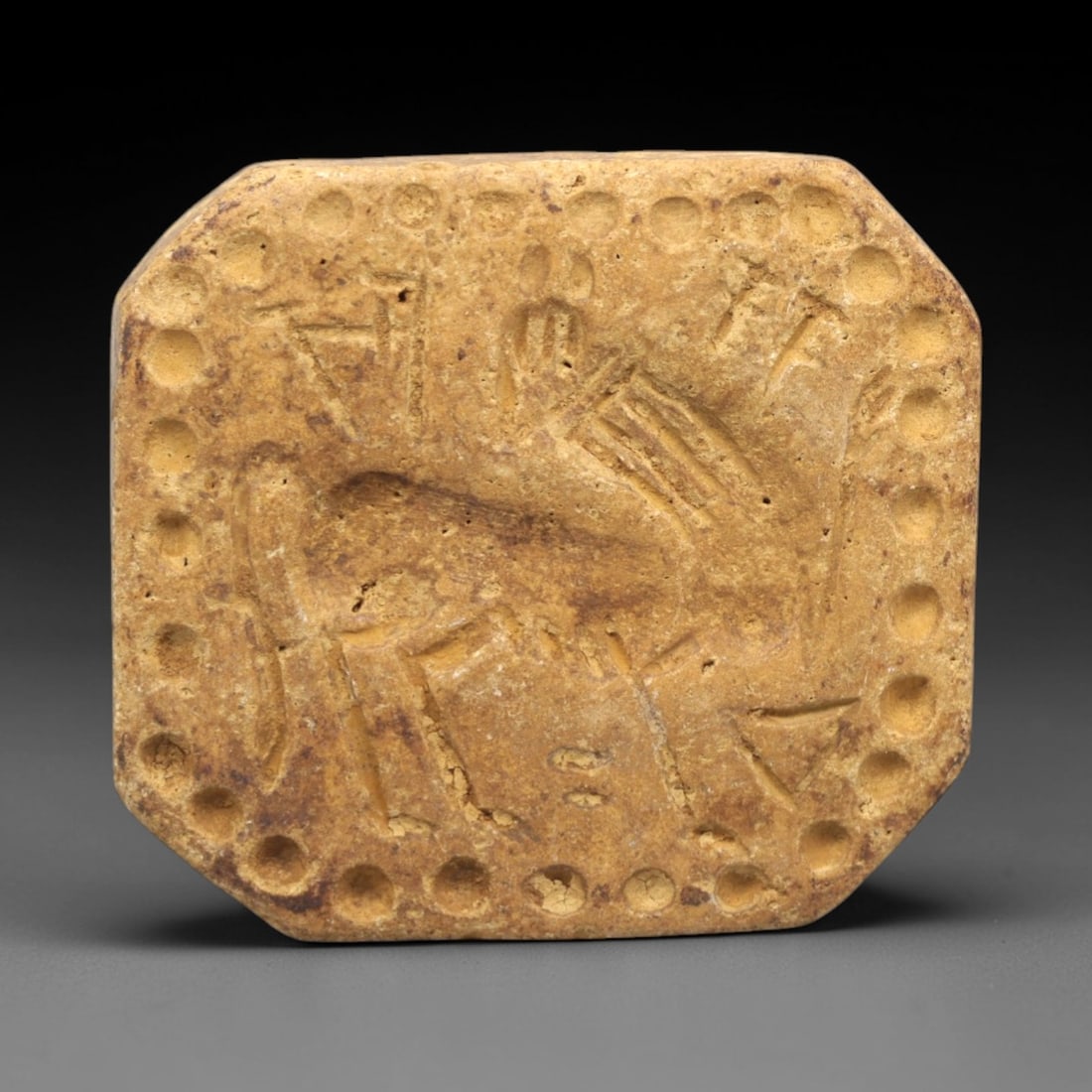 Ancient Stone Stamp Seal - Near Eastern Animal Amulet, 79ZP: Carved from warm tan stone, this octagonal stamp seal features a raised central animal motif in relief, surrounded by incised linear symbols and a border of evenly spaced circular perforations. The st