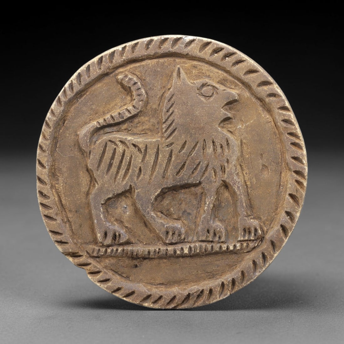 Ancient Steatite Plaque - Near Eastern Lion Relief, 92BE: Carved from steatite, the circular plaque features a raised relief of a striding lion, rendered with incised linear details defining its body, mane, and curled tail. The figure stands atop a decorated