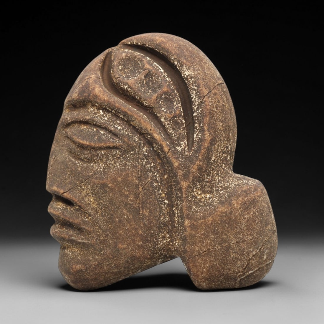 Ancient Stone Anthropomorphic Head - Near Eastern Idol, 86ZV: This artifact is a carved stone anthropomorphic head, executed in a granular, earth-toned stone consistent with ancient Near Eastern lithic traditions. The relief carving depicts a stylized profile fa
