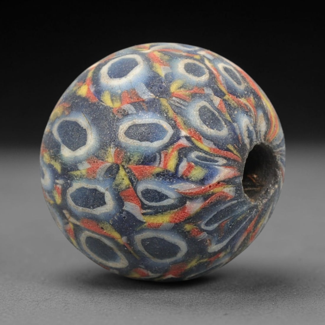 Ancient Mosaic Glass Bead - Roman Starburst Eye Amulet, 59WD: This spherical bead is crafted from core-formed mosaic glass, featuring a deep indigo blue base decorated with intricate concentric eye motifs surrounded by starburst-like radiating petals. Each eye m