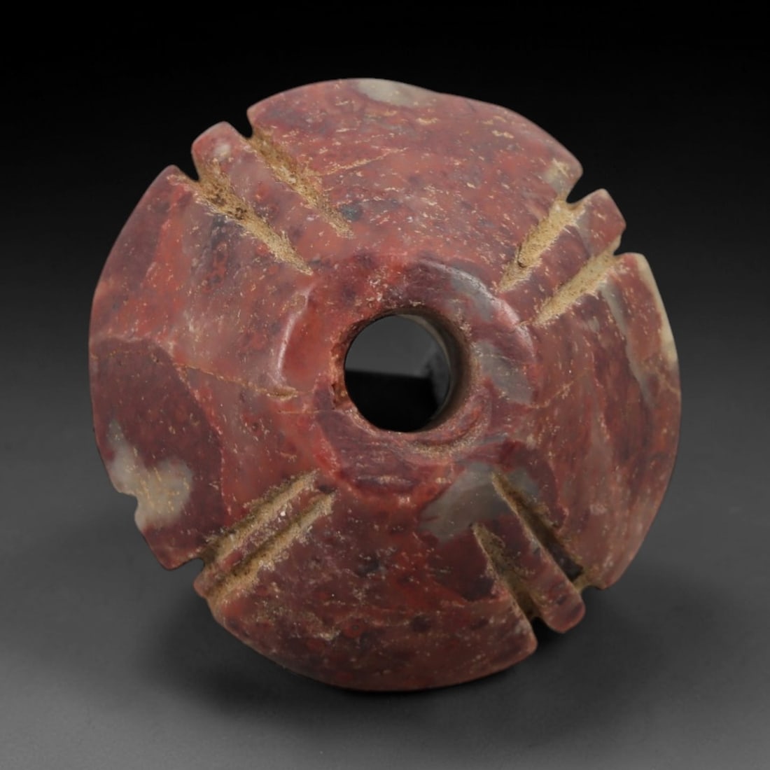 Ancient Stone Sun Amulet - Near Eastern Carved Pendant, 9AG: This amulet is carved from a speckled red stone with a weathered, earthy patina. The form is a circular disk with a central drilled hole, and four sets of paired incised linear grooves radiating from 