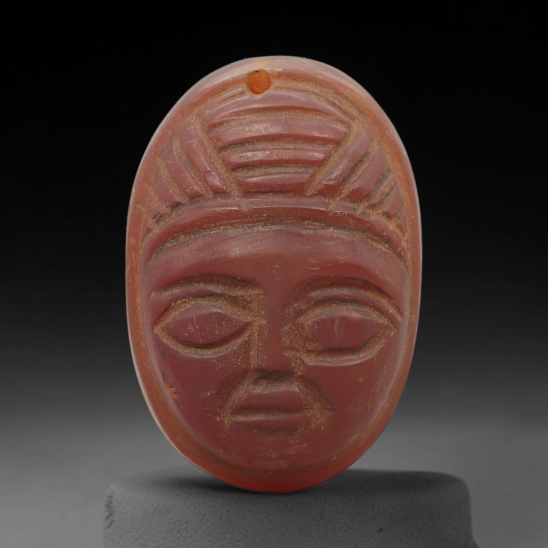 Ancient Carnelian Amulet - Near Eastern Mask Pendant, 39ZX: Carved from carnelian, the amulet is an oval plaque with a drilled suspension hole at the top, crafted in the ancient Near Eastern style. The front surface bears a raised relief portrait of a human ma