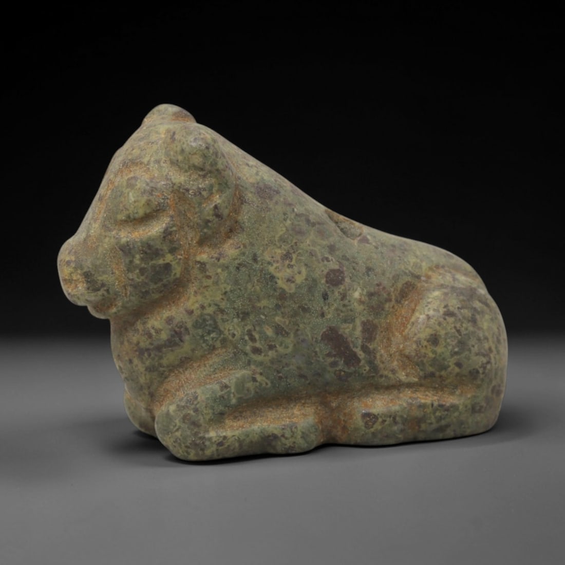 Ancient Stone Bovid Amulet - Near Eastern Animal Pendant, 24XQ: This amulet is carved from a mottled greenish-purple stone, shaped into a stylized reclining bovid form with a perforated suspension hole through the upper back, confirming its use as a wearable prote