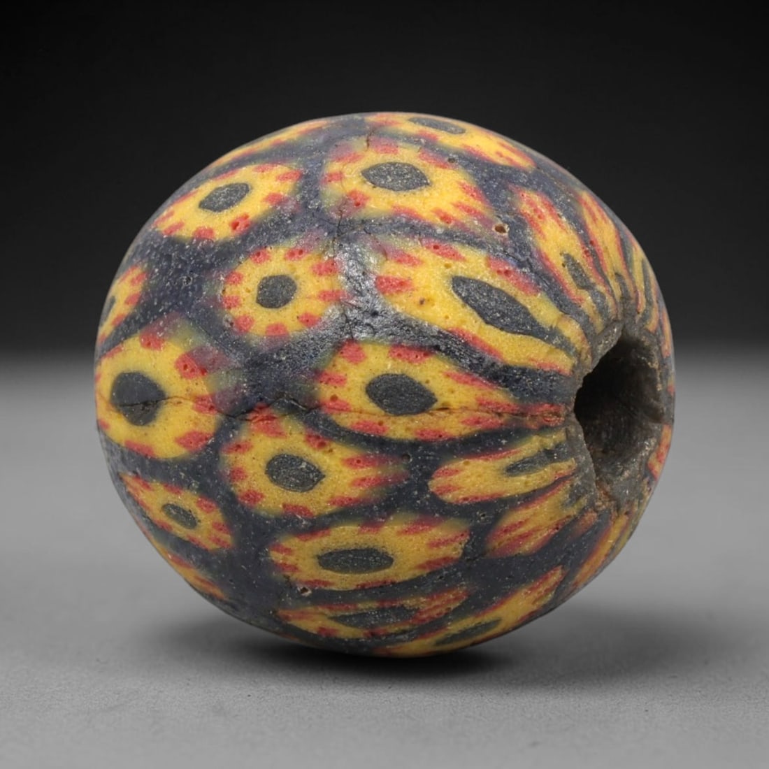 Ancient Mosaic Glass Bead - Roman Peacock Feather Amulet, 53WD: This spherical bead is crafted from core-formed mosaic glass, featuring a deep indigo base decorated with repeating peacock feather-inspired motifs. Each motif consists of a central dark blue inlay ou