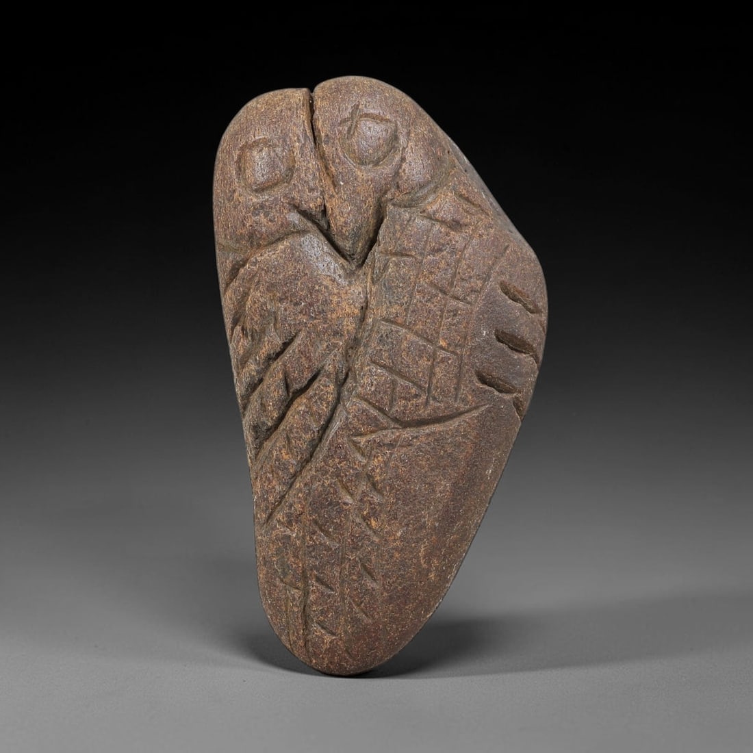 Ancient Stone Owl - Near Eastern Amulet Figurine, 49WJ: Carved from stone, this ancient Near Eastern amulet takes the form of a stylized owl, rendered with incised linear carvings that define the bird's facial features, wings, and body texture. The figure