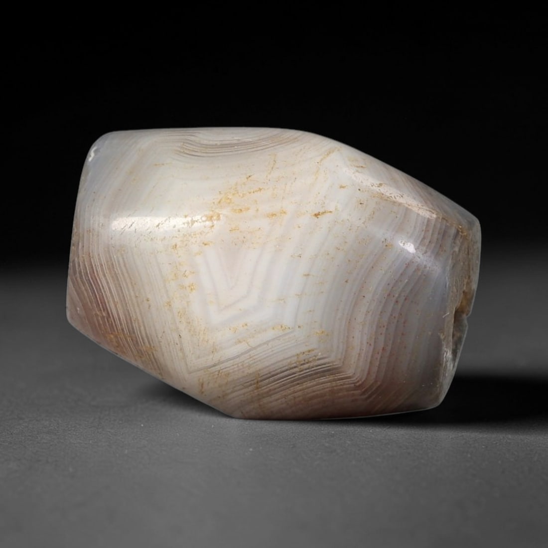 Ancient Agate Bead - Near Eastern Amulet, 51BC: This artifact is crafted from banded agate, showcasing striking concentric layered striations in pale grey, creamy white, and soft brown tones, shaped into a faceted barrel form through skilled ancien