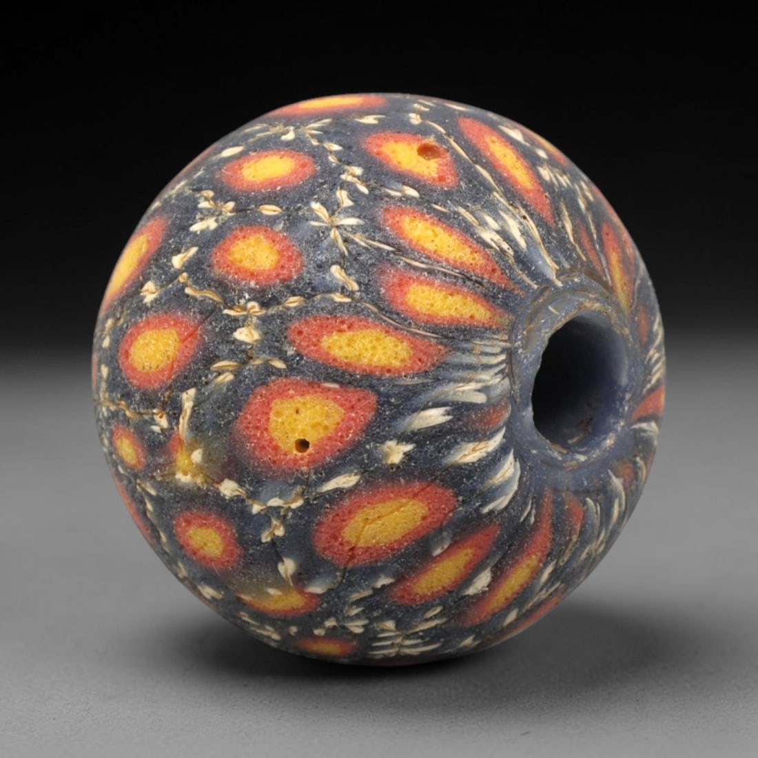 Ancient Mosaic Glass Bead - Roman Floral Pattern Amulet, 48WD: This spherical bead is crafted from core-formed mosaic glass, featuring a deep indigo blue base decorated with repeating teardrop-shaped floral motifs. Each motif consists of a central yellow glass in