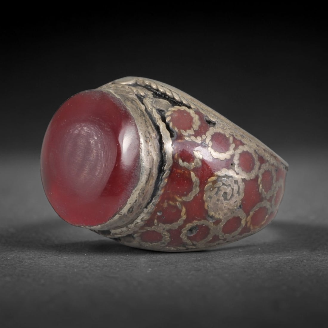 Antique Silvered Ring - Eurasian Steppe Carnelian Cabochon, 22BK: Silvered metal ring set with a large smooth carnelian cabochon, framed by twisted pseudo-filigree wirework. The band features a deep red-hued base with inlaid silver geometric and circular motifs, rai