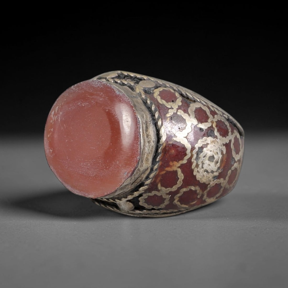 Antique Silvered Ring - Eurasian Steppe Carnelian Cabochon, 37BK: Silvered metal ring set with a large smooth carnelian cabochon, framed by twisted pseudo-filigree wirework. The band features a red enamel-like base with inlaid silver geometric and floral motifs, alo